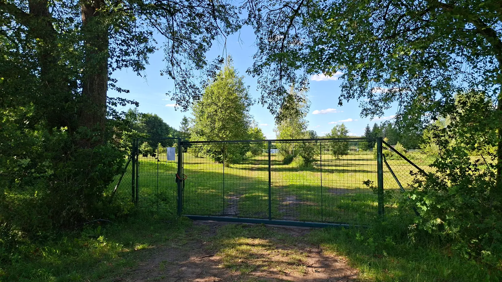 Zdjęcie o orientacji poziomej ukazuje zamkniętą, zieloną bramę wykonaną z metalowej siatki, która stanowi wejście na otwarty, słoneczny teren zielony. Kompozycja zdjęcia jest symetryczna, gdzie brama znajduje się na środku kadru, otoczona po obu stronach gęstymi, zacienionymi drzewami, które tworzą naturalną ramę dla widoku. Przez oczka siatki widać rozległą polanę z drzewami i krzewami w świetle letniego, bezchmurnego popołudnia. Na lewym skrzydle bramy przymocowana jest biała kartka z informacją, która jest jednak nieczytelna z tej odległości. Poniżej znajduje się treść widniejąca na tabliczce: "UWAGA! Teren prywatny Wstęp wzbroniony". Całość utrzymana jest w żywej, letniej kolorystyce z wyraźnym kontrastem między ciemnym cieniem drzew na pierwszym planie a jasną, nasłonecznioną trawą w głębi.